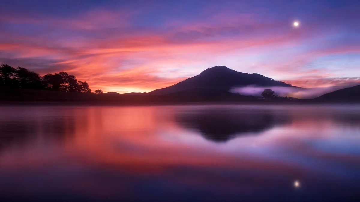 Golden hues of sunset dance across the mountain peaks, mirrored perfectly in the tranquil lake's glassy surface. A dramatic…