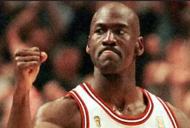 a close-up photo of basketball player Michael Jordan in a white Chicago Bulls jersey with red trim, raising his right fist…