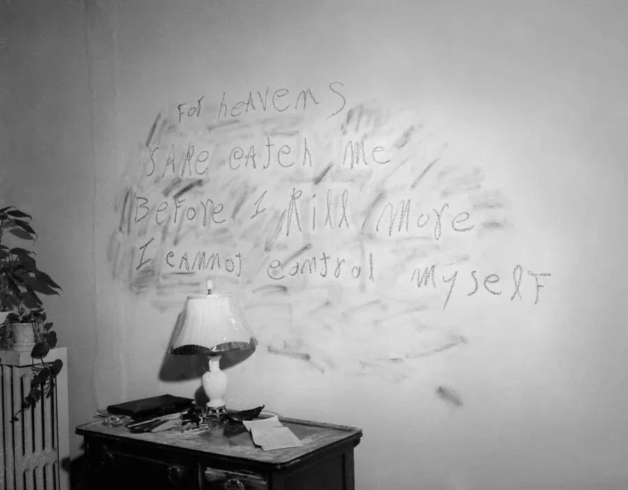 A black-and-white photo shows a messy room with a lamp on a cluttered dresser and a potted plant nearby. On the wall behind…