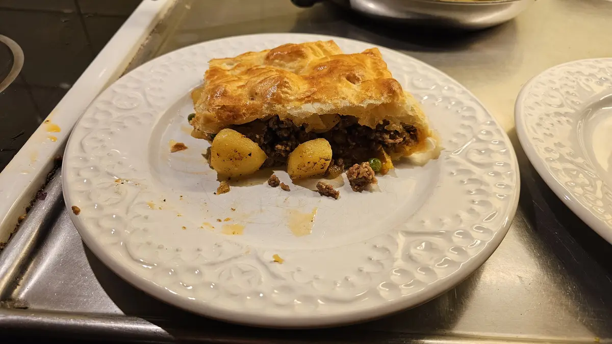 A partially eaten slice of meat pie with golden-brown pastry sits on a white decorative plate, revealing a filling of ground…