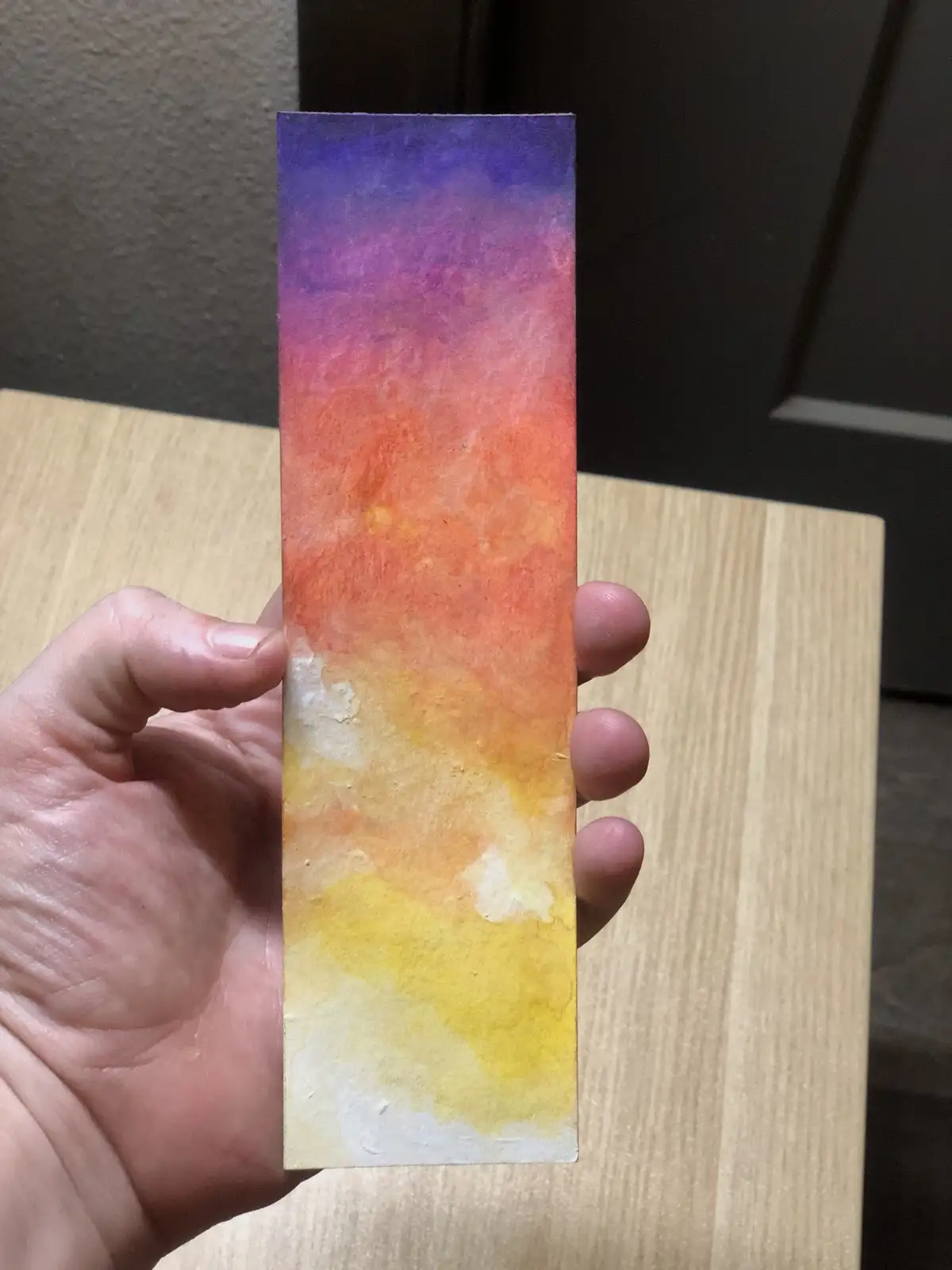 A hand holds up a vertically oriented rectangular piece of paper with a watercolor gradient transitioning from purple at the…