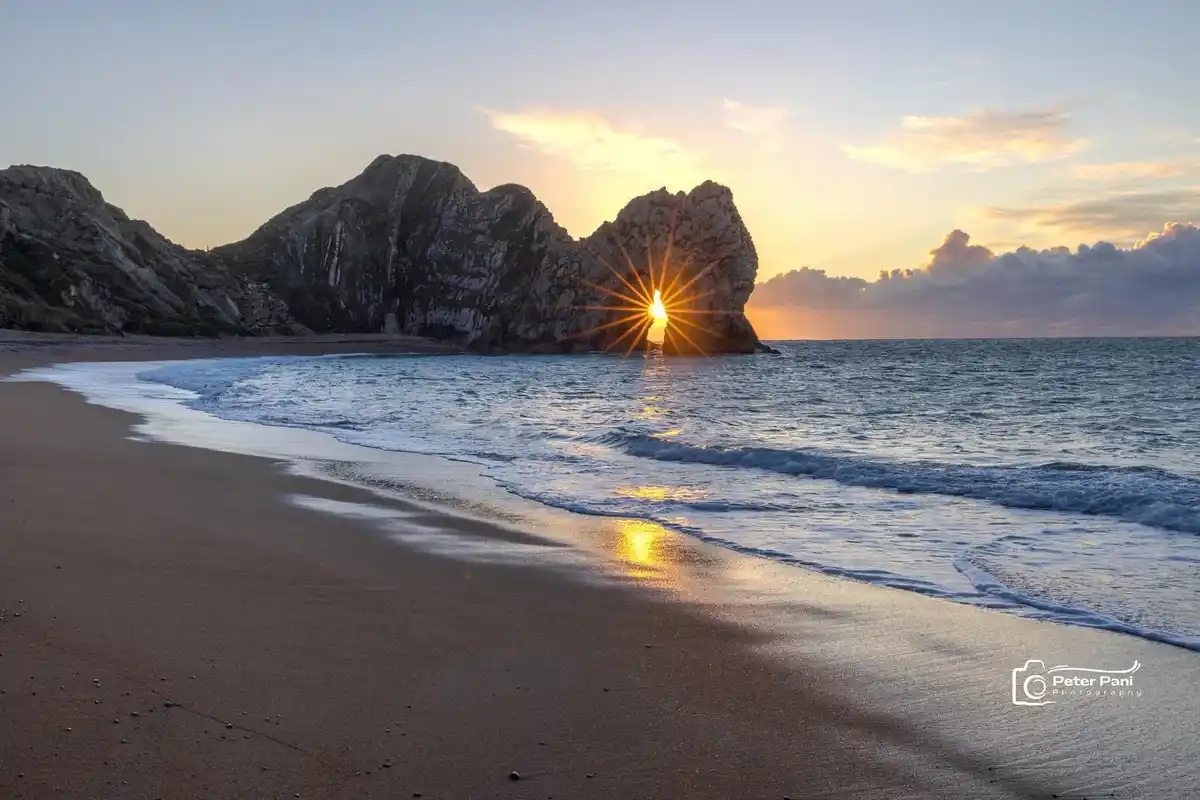 a serene sunset photograph shows the sun shining through a natural hole in a large coastal rock formation, creating a bright…