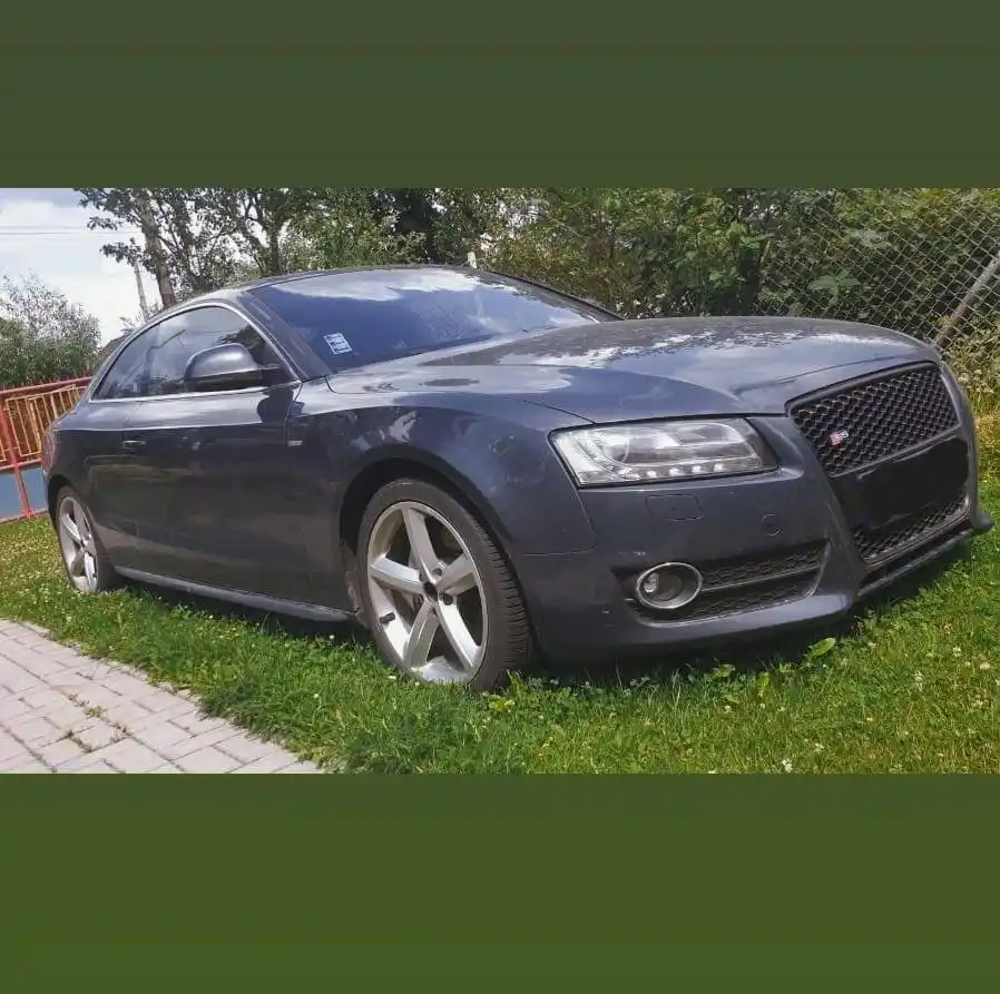 A dark gray Audi S5 coupe is parked crookedly on grass next to a paved walkway, with trees and a chain-link fence in the…