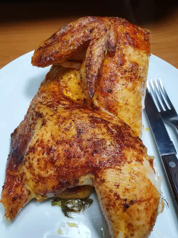 A close-up photo shows a roasted chicken on a white plate, with its wings folded up over its back, next to a knife and fork…