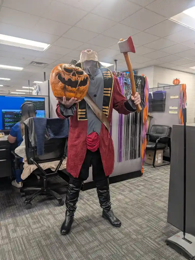 A person in an office wearing a pirate costume with a severed pumpkin head for a head, holding the pumpkin in one hand and…