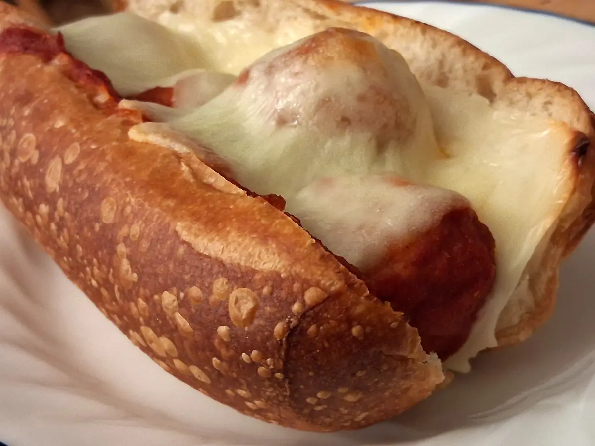 A close-up photo of a hot dog in a pretzel-style bun, topped with melted cheese and red sauce, resembling a pizza. The text…