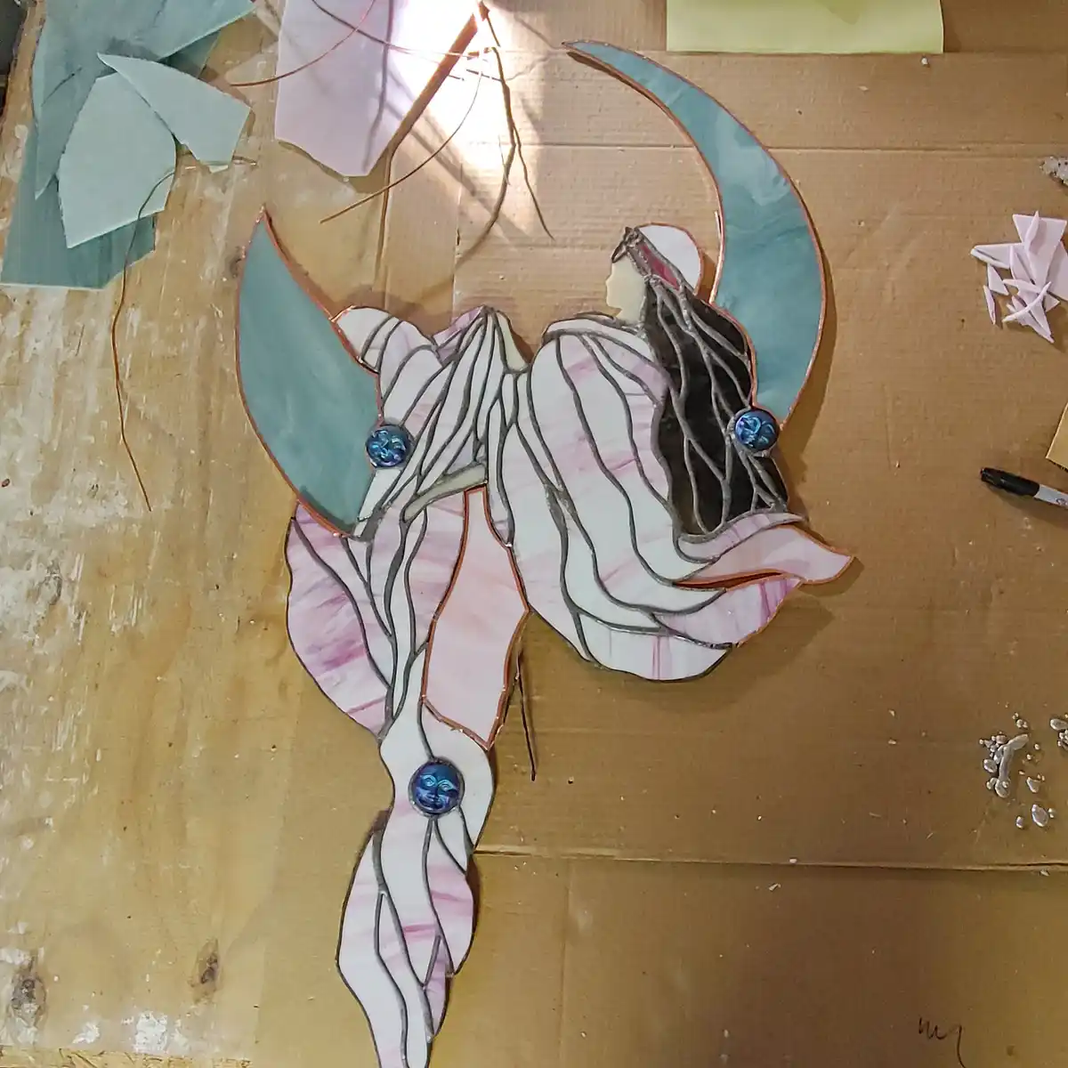 A stained glass artwork of a stylized female figure with flowing pink and white robes, set against a crescent moon backdrop…