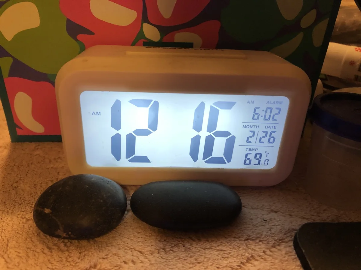 A digital alarm clock displays 12:16 AM, with additional info showing an alarm set for 6:02 AM, date 2/26, and temperature…