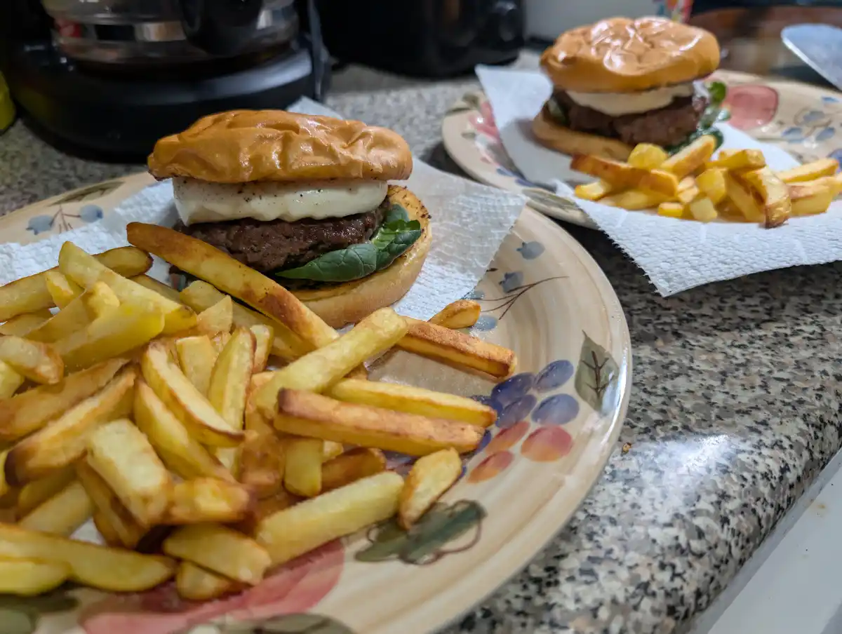 Two plates of homemade burgers with melted cheese, spinach, and fries sit on a granite countertop. The burgers are on…