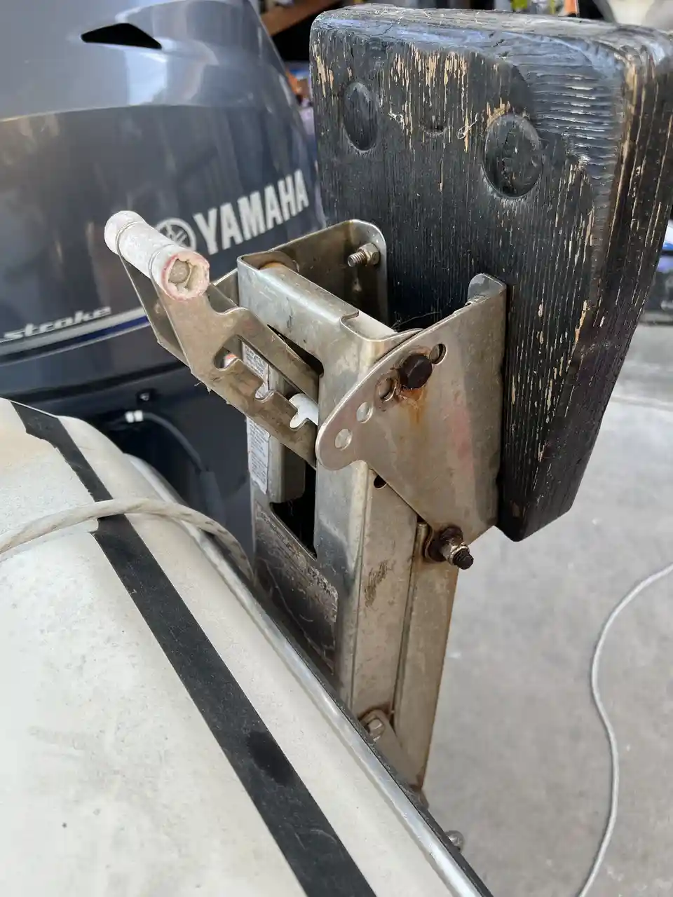 A close-up photo shows a weathered, black-painted wooden post with a rusty metal bracket attached, holding a Yamaha outboard…