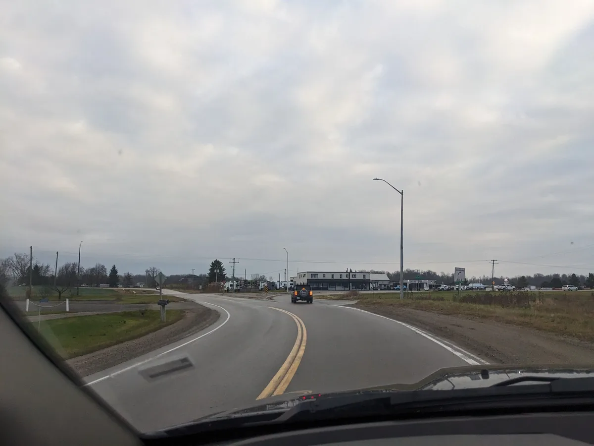 A photo taken from inside a car shows a road curving toward a commercial area with buildings and parked vehicles under an…