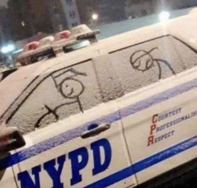 A snowy NYPD police car has stick figures drawn on its rear window: one smiling officer driving and one frowning passenger…