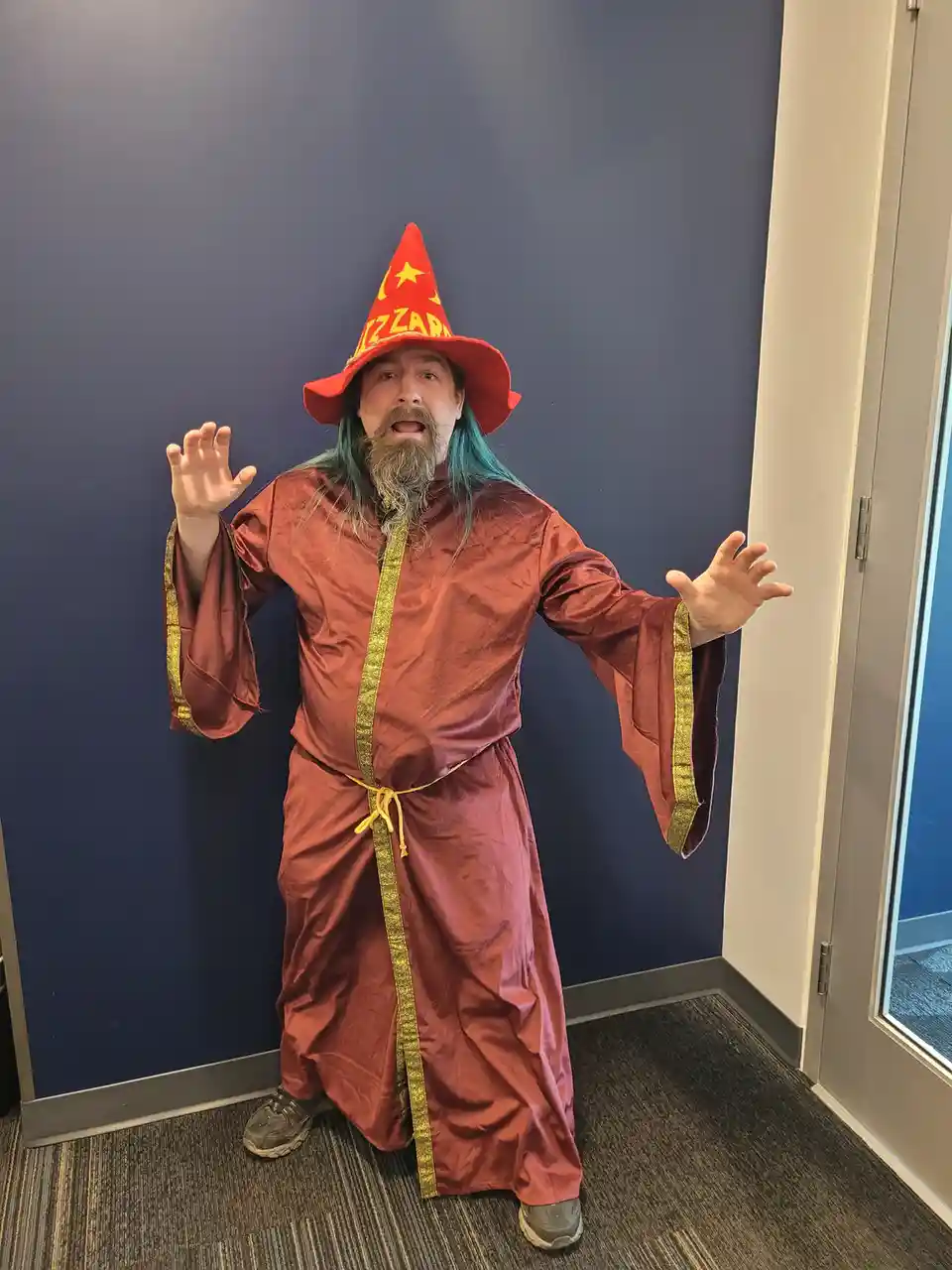 A man in an office setting wears a red wizard hat with 'WIZARD' written on it and a red robe with gold trim. He has a long…