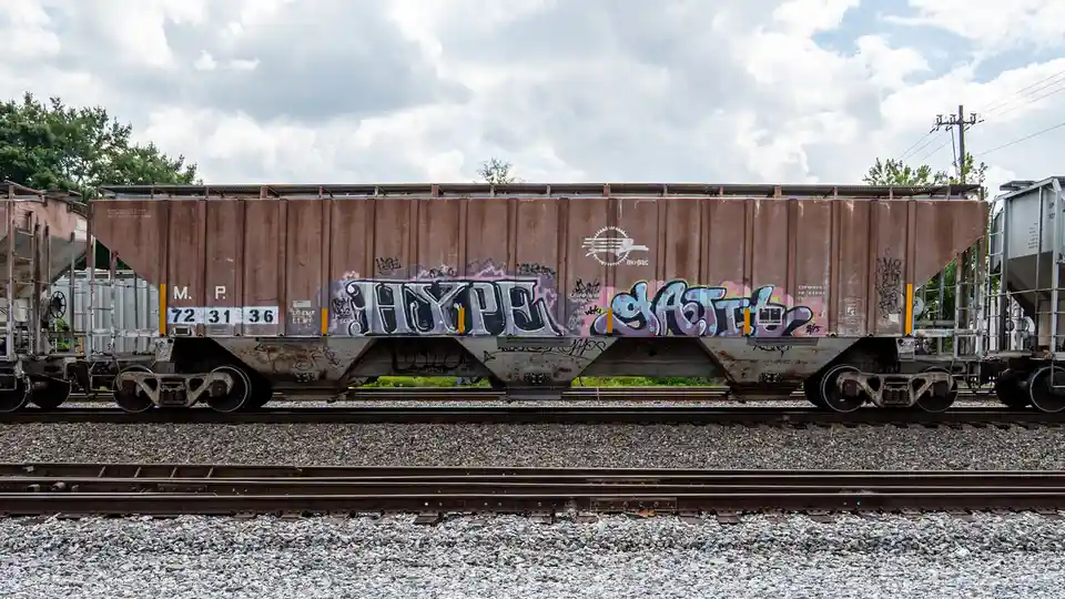 A rusty brown freight train car covered in colorful graffiti sits on railway tracks under a cloudy sky. The most prominent…