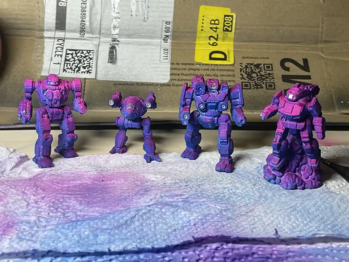 Four purple-painted robot action figures stand in a row on a paper towel with purple and blue paint stains. The background…
