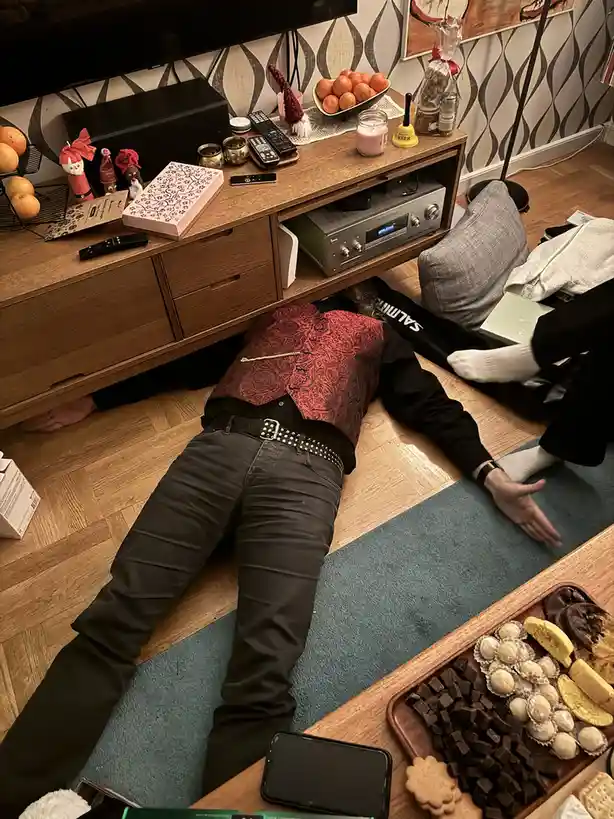 A person is lying motionless on a wooden floor under a TV stand, wearing a red patterned vest, black shirt, and dark pants…