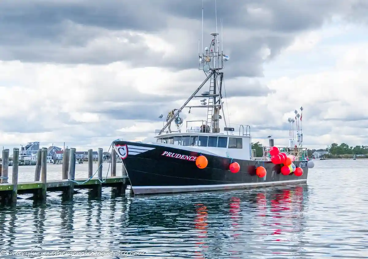 A black and white fishing boat named 'PRUDENCE' is docked at a wooden pier under a cloudy sky. The boat has orange buoys and…