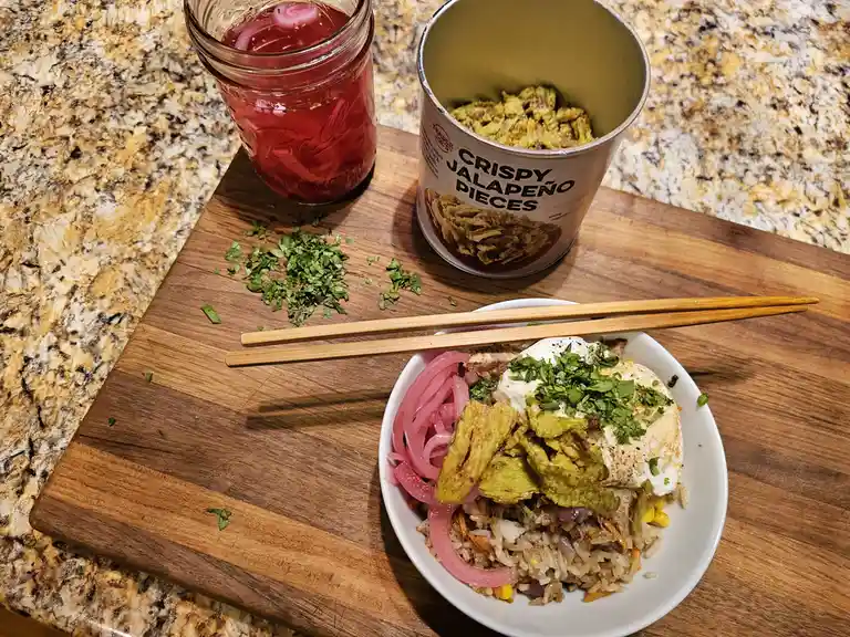 A bowl of rice with meat, pickled red onions, a fried egg, and chopped cilantro sits on a wooden cutting board with…