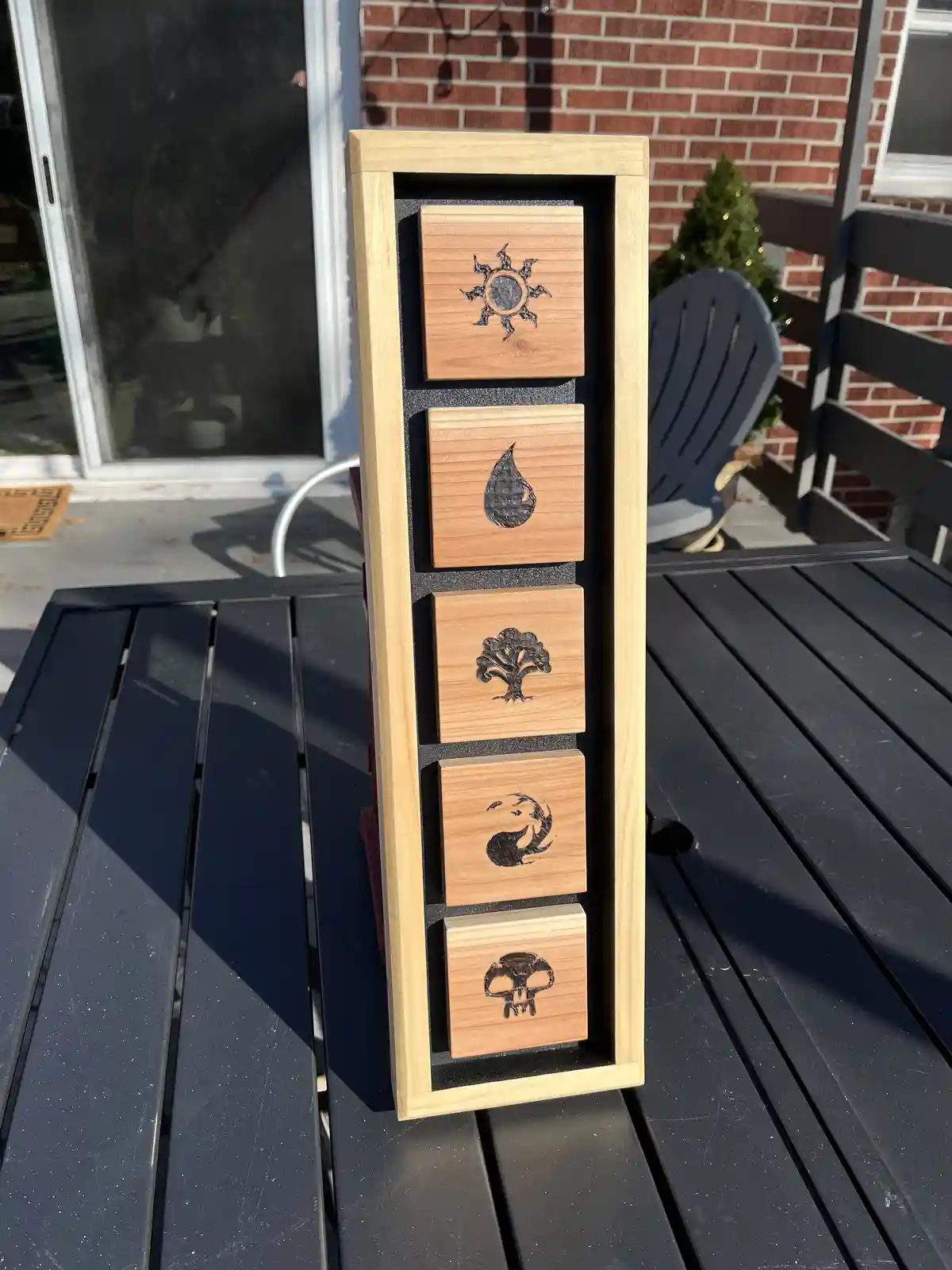 A vertical wooden display case holds five square wooden blocks, each engraved with a symbol: a sun, a water droplet, a tree…