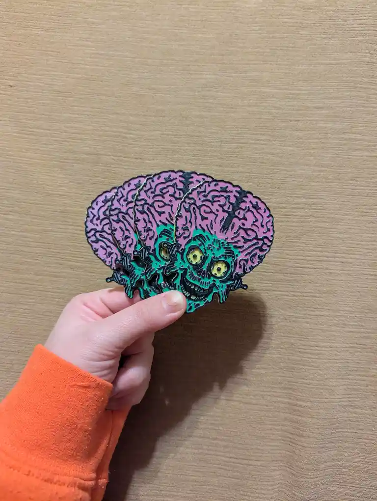 A hand holds a stylized, layered enamel pin depicting a pink brain with a green, skull-faced creature emerging from its…
