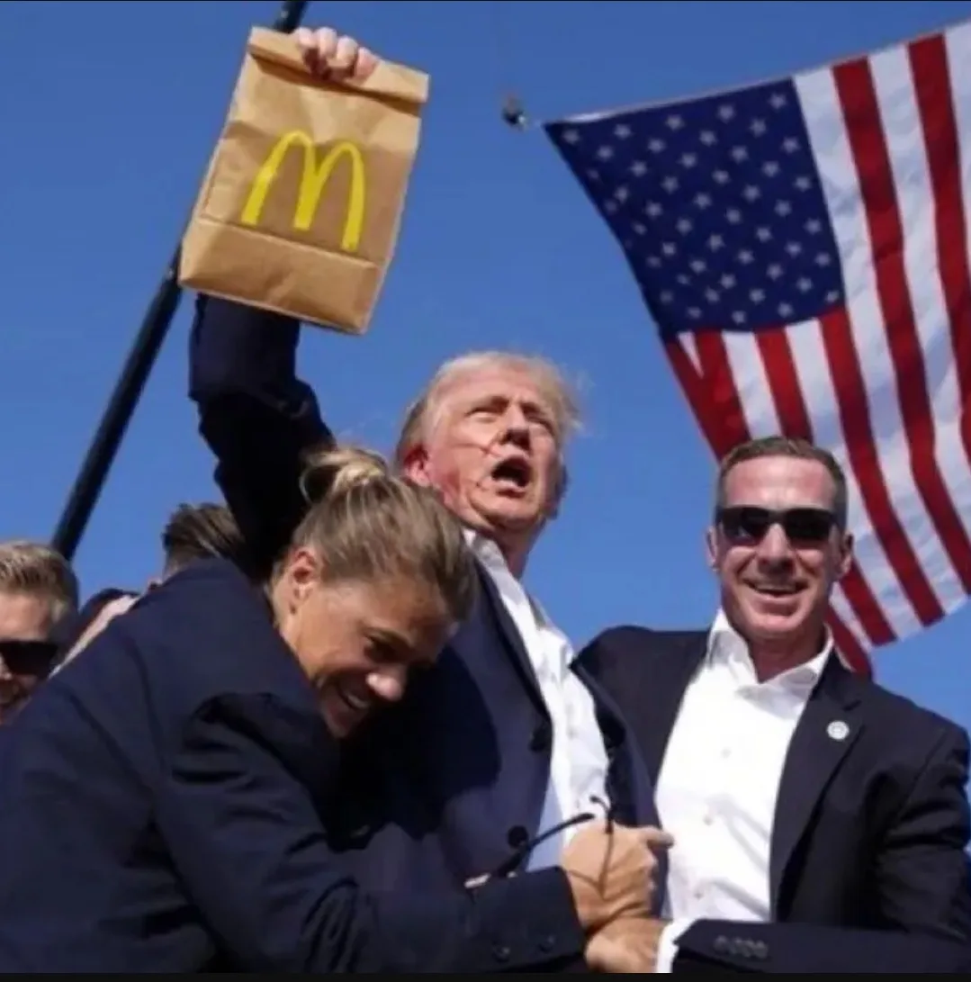 A man in a dark suit holds up a McDonald's paper bag with the golden arches logo against a clear blue sky, with an American…
