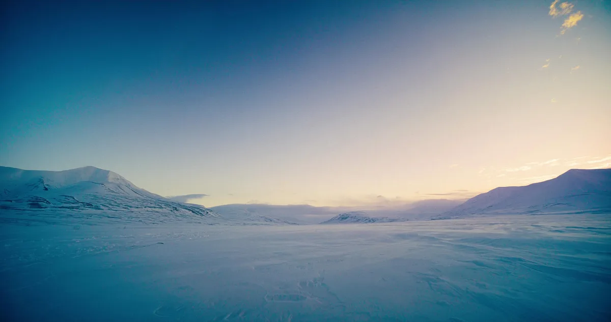 A wide, serene landscape of snow-covered mountains under a gradient sky transitioning from deep blue to soft orange near the…