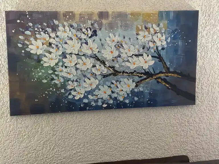 A painting of a flowering branch with white blossoms and orange centers hangs on a textured wall. The background is a blend…