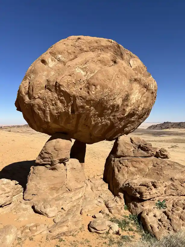 A large, rounded boulder is precariously balanced on two smaller rock pillars in a desert landscape under a clear blue sky…
