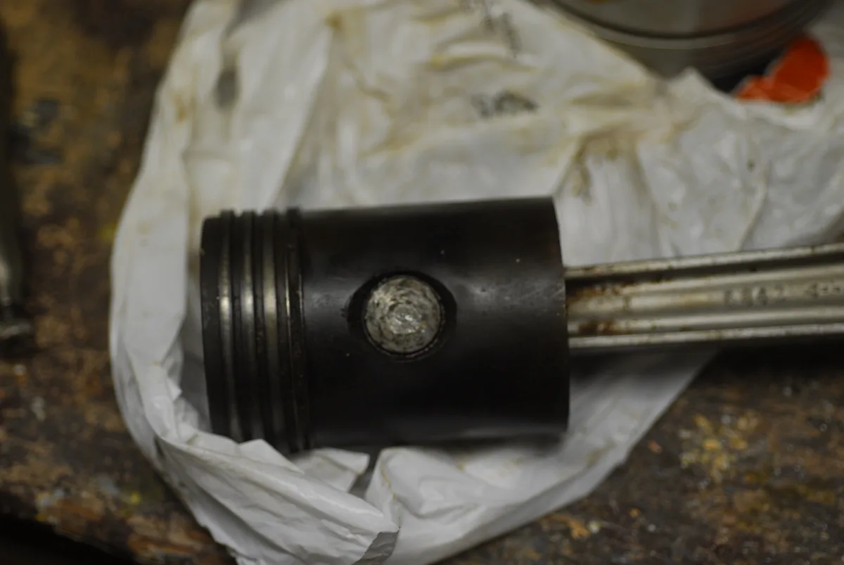 A close-up photo shows a dirty, used piston resting on a crumpled white paper towel on a rusty workbench. The piston has…