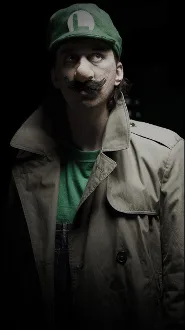 A man with a mustache, wearing a green cap with an 'L' logo and a green shirt under a beige trench coat, stares seriously at…