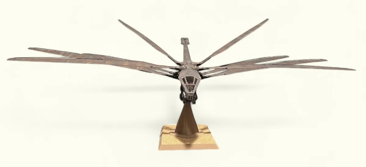 a detailed metal sculpture of a dragonfly with long, segmented wings and a segmented body, mounted on a textured brown…