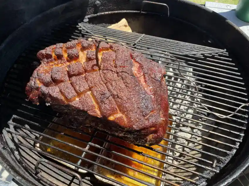 a large, heavily seasoned piece of meat with a grid pattern of cuts sits on a black charcoal grill, showing a dark, crispy…