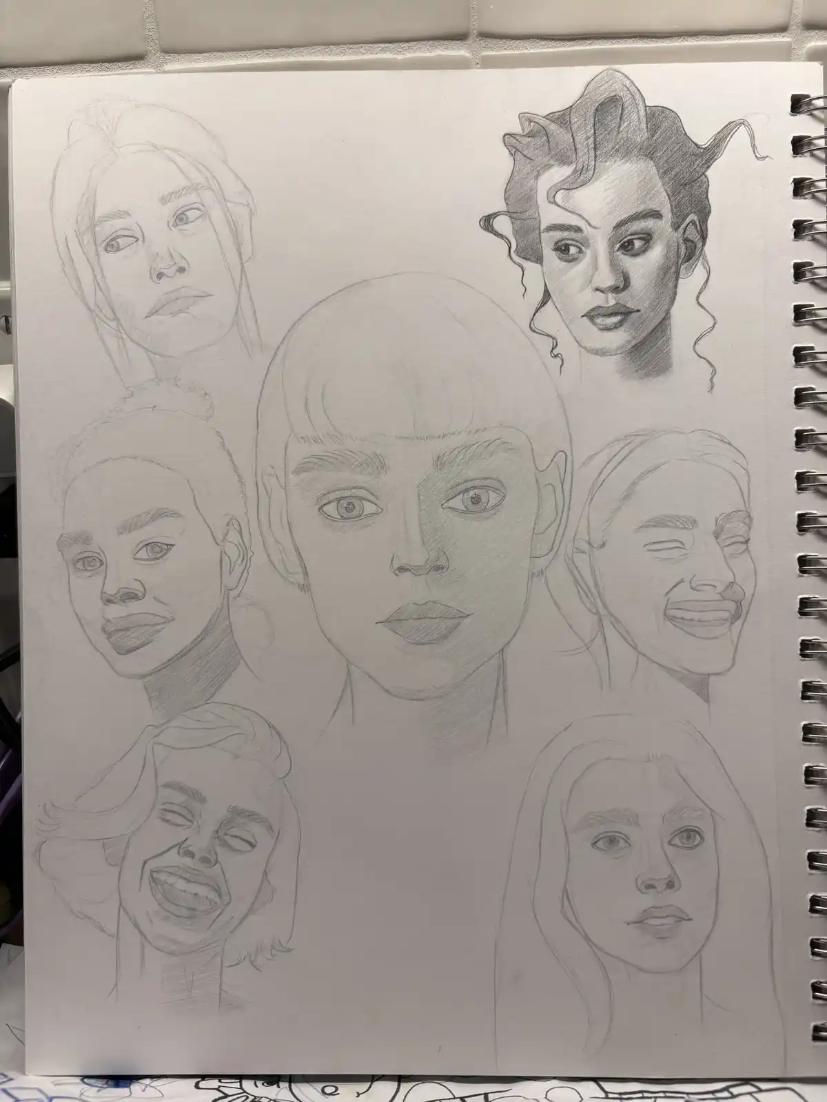 A pencil sketch in a spiral-bound sketchbook shows seven different female faces drawn in various styles and expressions…