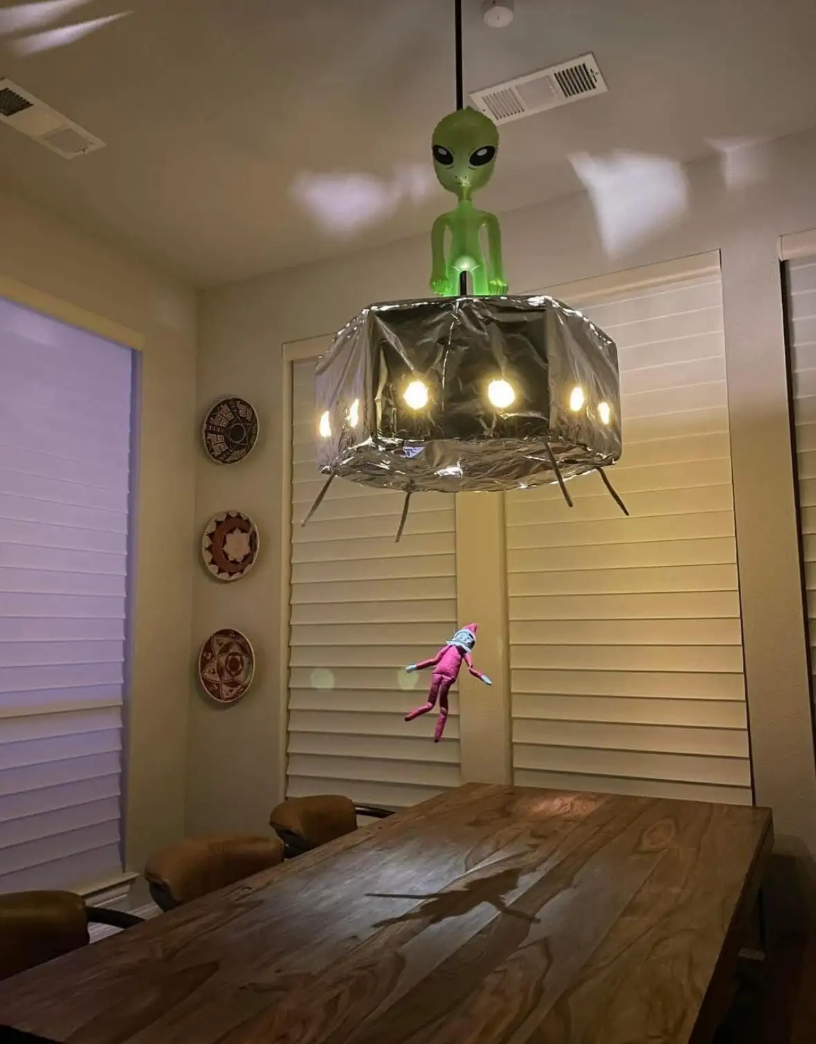 A dining room features a chandelier modified to look like an alien spaceship, with a green alien figurine on top and…