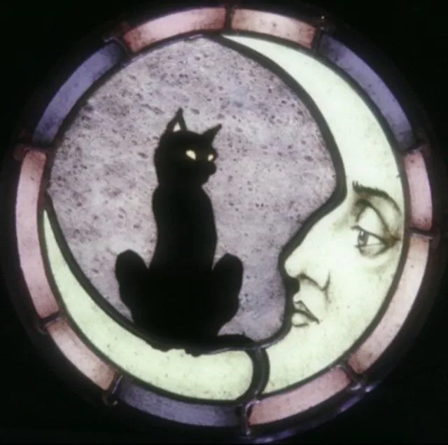 a stained glass window depicts a black cat with glowing eyes sitting atop a crescent moon, which has a human face looking…