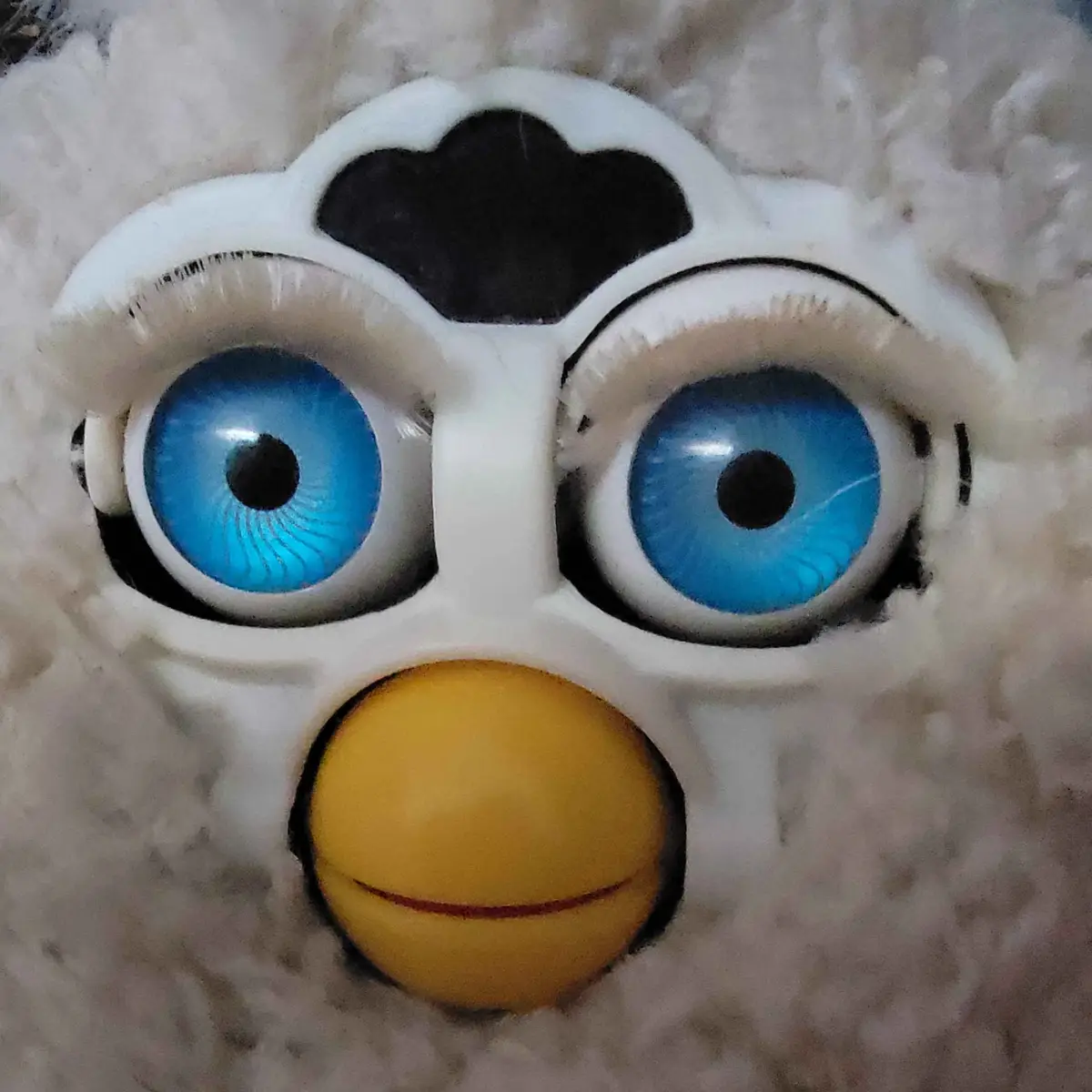 A close-up of a white, fluffy Furby toy with large, bright blue eyes and a yellow beak-like nose. The Furby has a black…