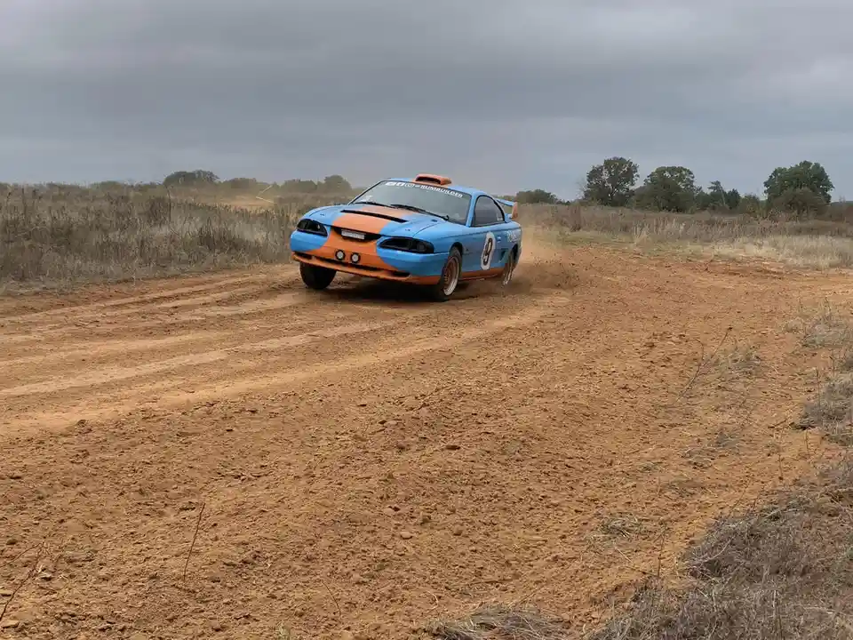 A blue and orange rally car with the number 9 on its side is speeding along a dirt road, kicking up dust under an overcast…