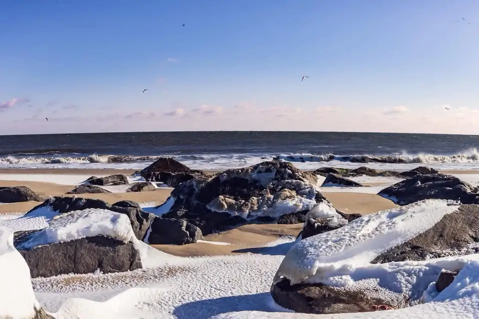 A snowy beach scene with large rocks covered in snow, waves crashing in the background under a clear blue sky. The overlaid…