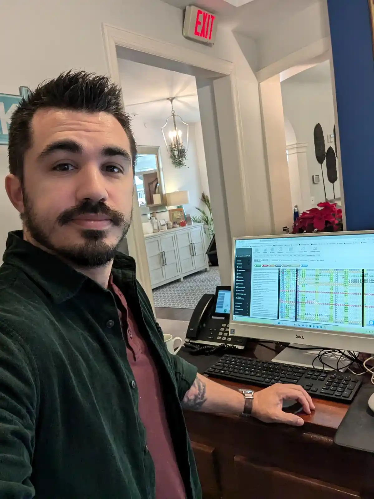 A man with a beard and dark hair takes a selfie at a wooden desk, looking directly at the camera with a slight smirk. Behind…