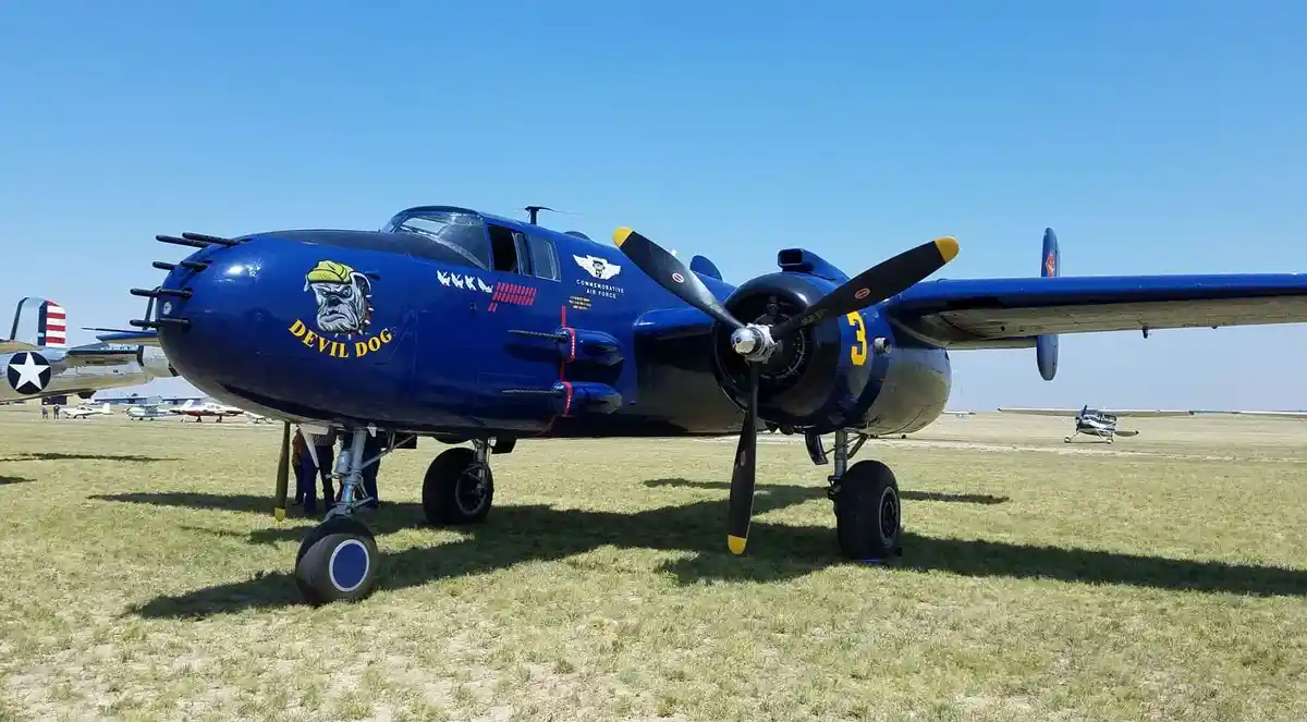 A blue vintage military aircraft, likely a WWII-era bomber, is parked on a grassy airfield under a clear blue sky. The…