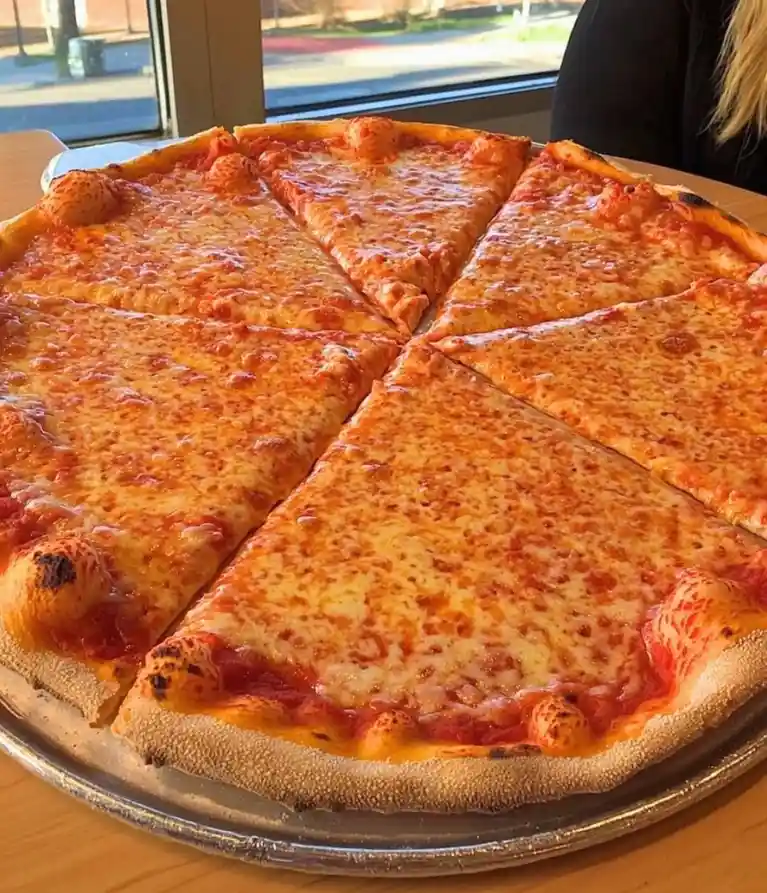 A close-up photo of a large, cheesy cheese pizza on a metal tray, sliced into triangular pieces with a thick, slightly…