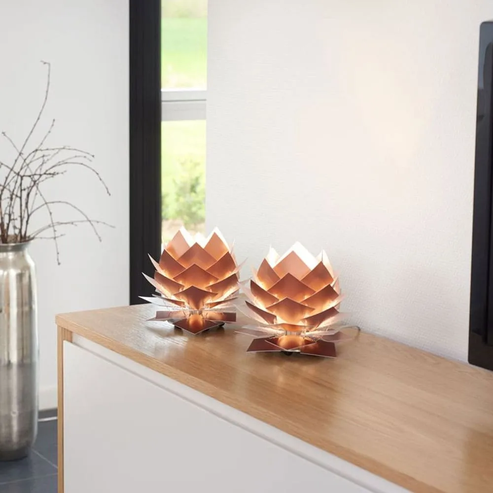 Two copper-colored, artichoke-shaped table lamps with layered petal-like structures sit on a light wood cabinet in a modern…