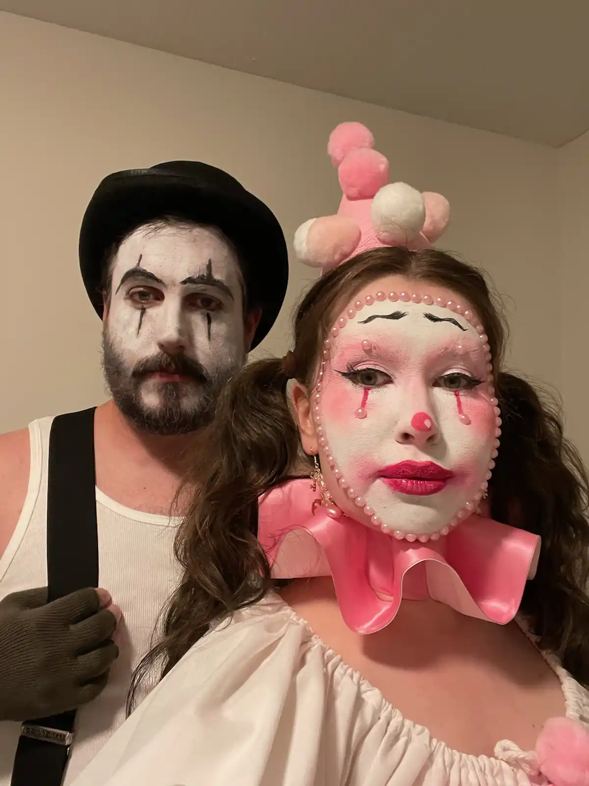 A man and woman dressed as clowns pose for a photo. The man has white face paint, black tear streaks, and a black bowler…