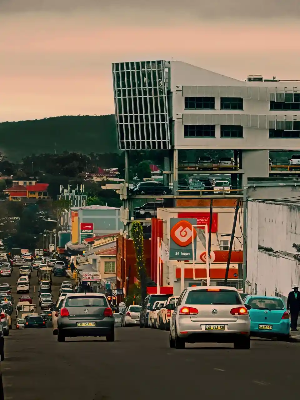 A busy urban street scene at dusk with heavy traffic, cars lined up, and a modern building with a parking garage. Overlaid…
