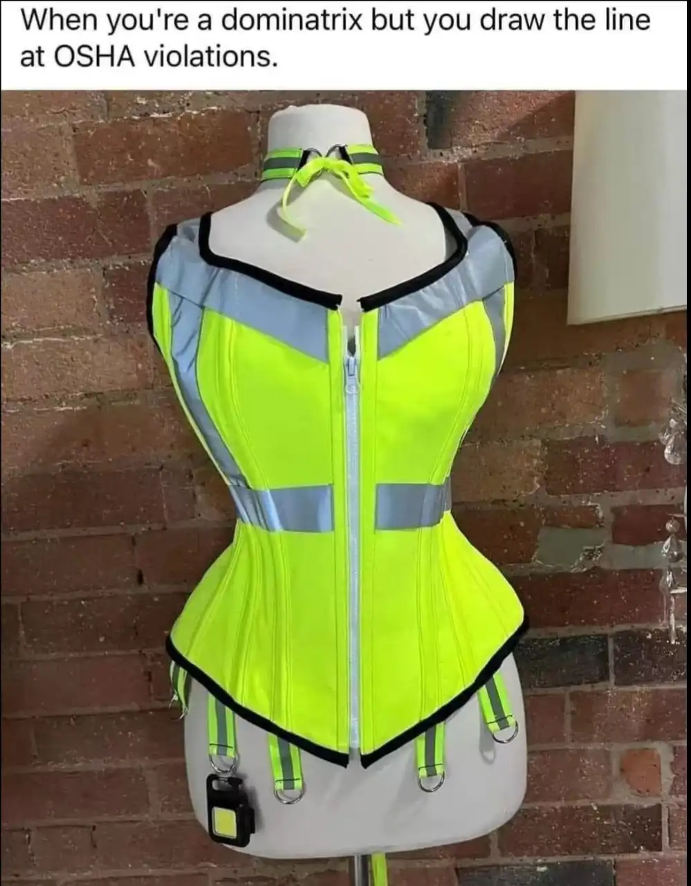 A mannequin wears a bright yellow safety vest with black trim and reflective stripes, modified into a corset-like garment…