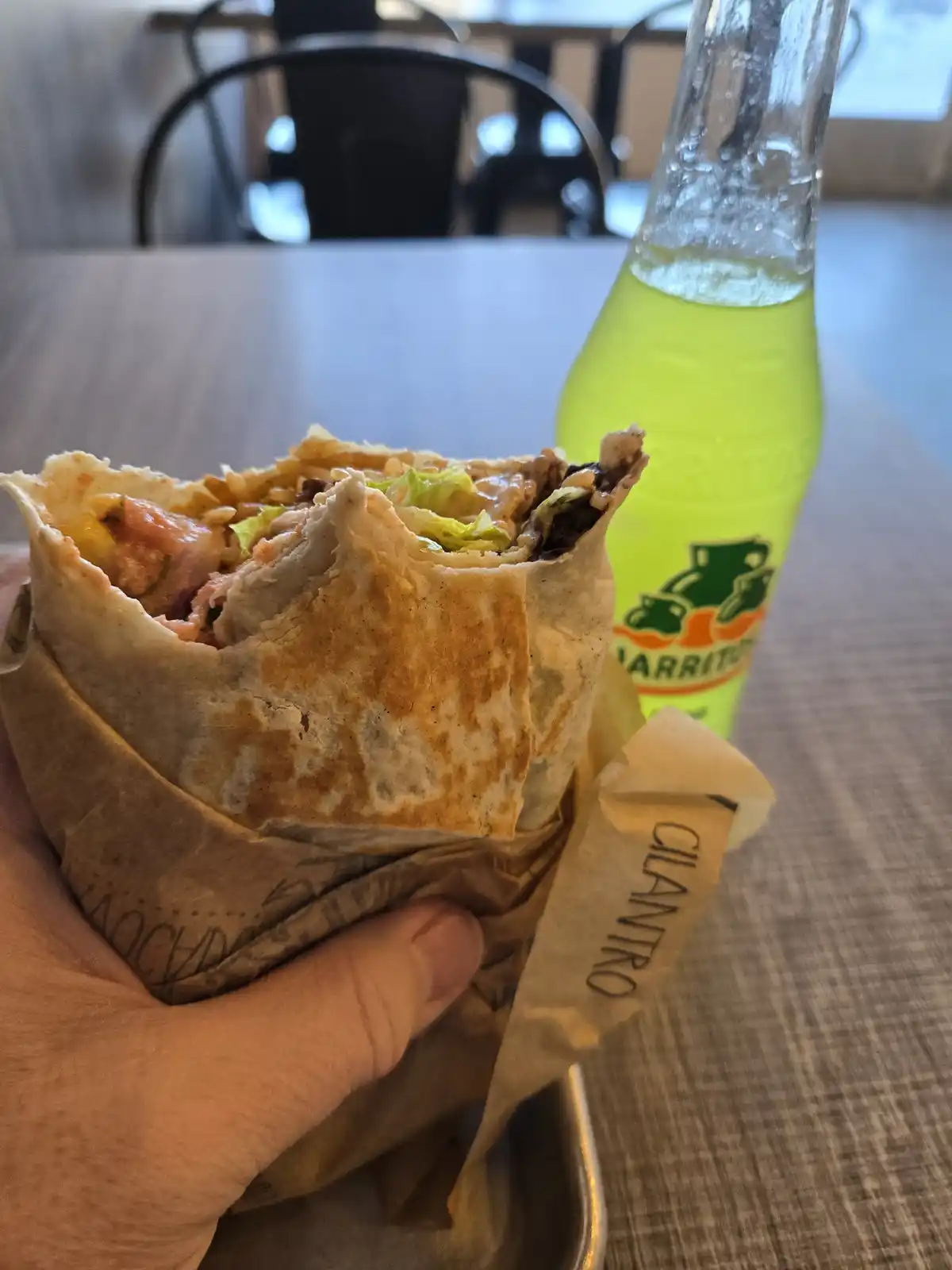 A hand holds a partially eaten burrito wrapped in brown paper with 'CILANTRO' printed on the wrapper, next to a green bottle…
