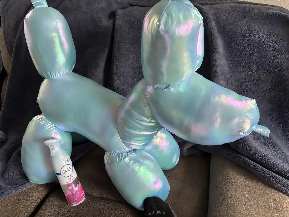 Two iridescent, balloon-style dog-shaped pillows sit side-by-side on a dark gray blanket over a brown couch. A bottle of…