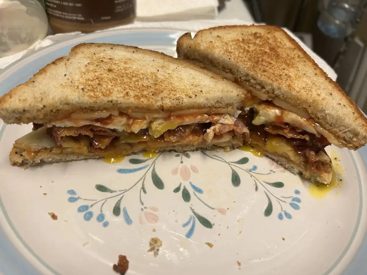 A close-up photo shows a grilled cheese sandwich cut in half, revealing melted cheese, bacon, and egg yolk oozing out onto a…