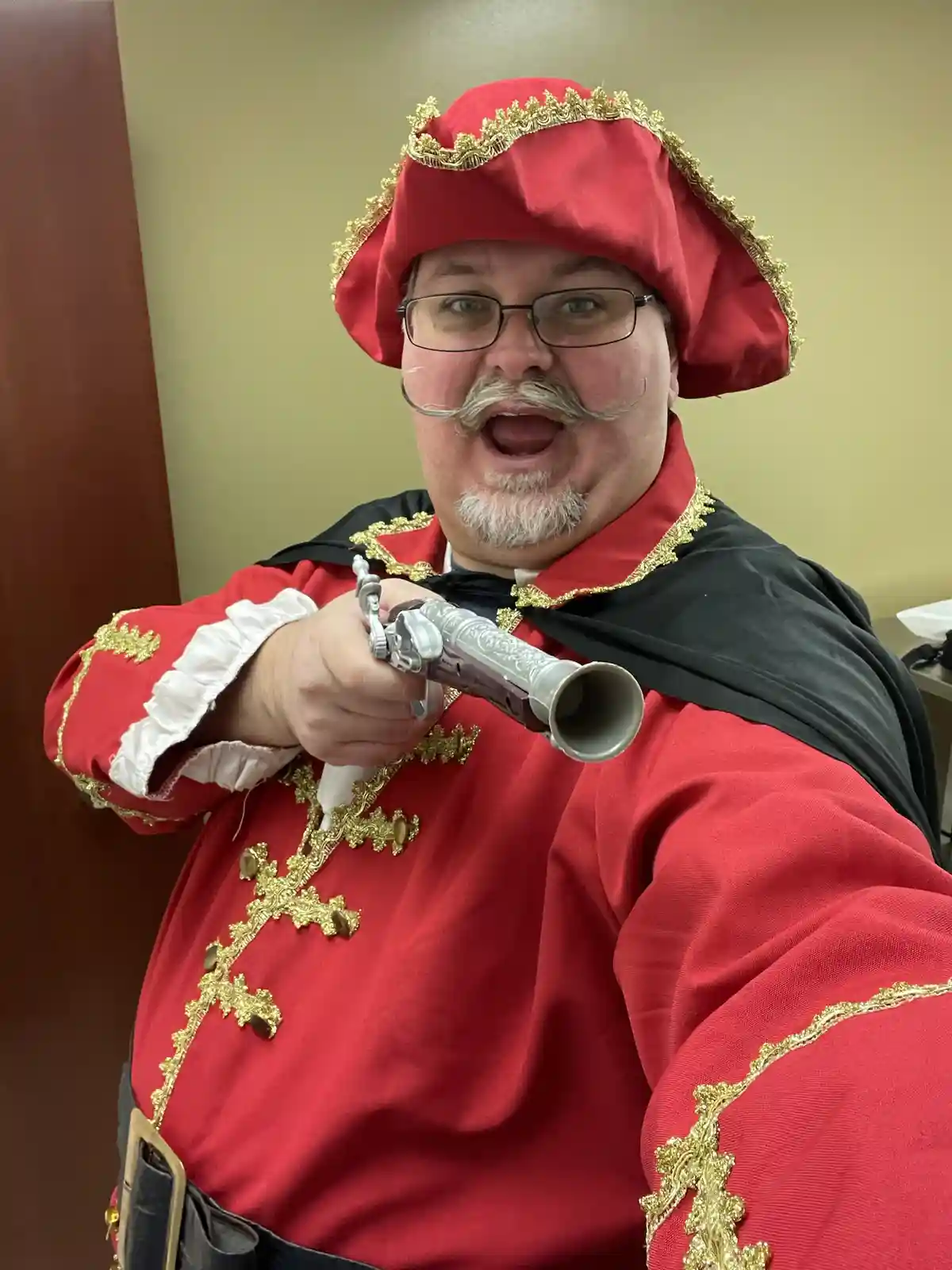 A man in a red and gold pirate costume with a fake mustache and glasses holds a toy pistol pointed forward, mouth open as if…