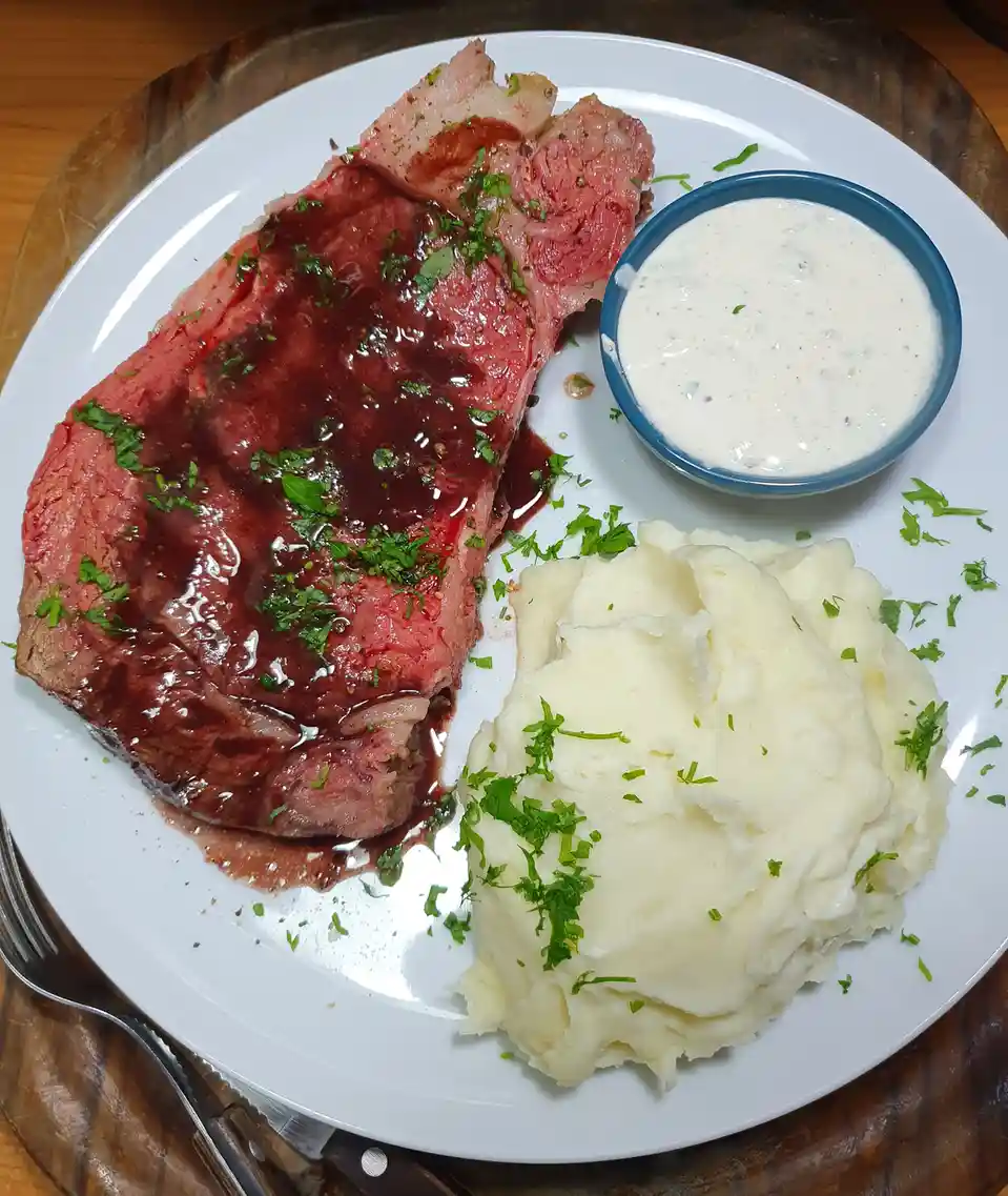 A plate of food featuring a large, rare-cut steak with a dark sauce and chopped green herbs, served alongside a mound of…